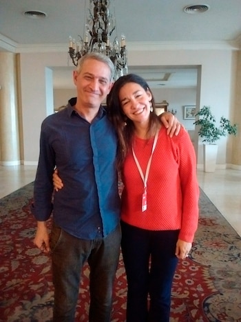 Hombre de camisa azul y pantalón oscuro, con una mujer de jersey rojo, pantalón oscuro y acreditación. Ambos sonríen en el interior de un salón con alfombra y candelabro