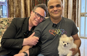 Rafi, con un pin de corazón arcoíris en la solapa, sonríe mientras Ashraf besa a un perro pomerania blanco, sentados en un sofá estampado (Fuente Latina)