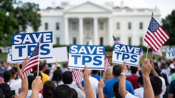 Una multitud de personas levanta carteles azules con la leyenda "SAVE TPS" y banderas de Estados Unidos frente a un edificio blanco con columnas.
