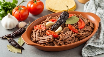 Plato con carne desmenuzada, tomate, chiles secos, ajo y laurel. Alrededor hay tortillas, tomates frescos, cabeza de ajo, cilantro y un paño.
