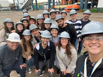 Un grupo grande de personas sonríe a la cámara. Todos visten cascos grises y gafas de seguridad. Al fondo, se observan edificios y maquinaria industrial