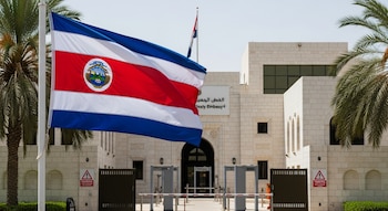 Bandera de Costa Rica ondeando en primer plano frente a la fachada de una embajada genérica con arquitectura de piedra clara, palmeras y controles de seguridad.