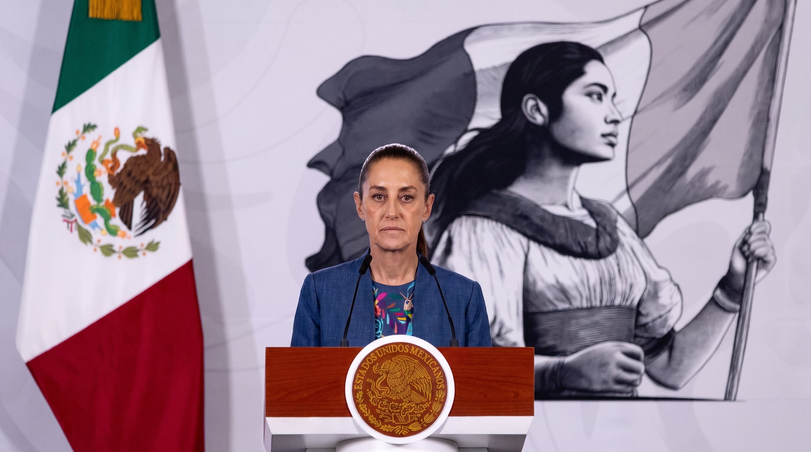 La funcionaria Claudia Sheinbaum se dirige a la audiencia desde un podio, acompañada por la bandera nacional y una representación artística de mujer con bandera. (Imagen Ilustrativa Infobae)