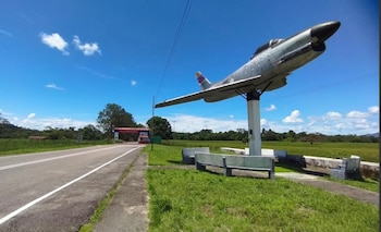 Un avión militar sobre un pedestal blanco, bancos de cemento, una carretera asfaltada, árboles y campos de vegetación