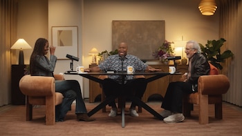 Michelle Obama, un hombre y Steven Spielberg sentados en sillas de terciopelo naranja alrededor de una mesa negra, con micrófonos y tazas, en un estudio de podcast