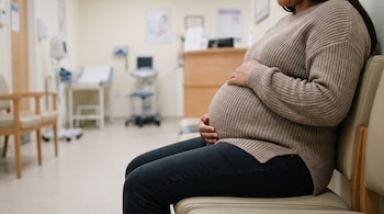 Una mujer embarazada con un suéter beige y pantalones oscuros sentada en una silla clara en una clínica con equipo médico en el fondo.