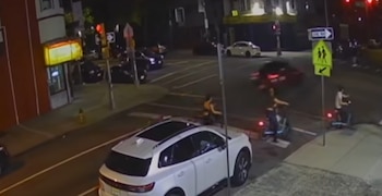 Vista aérea de un cruce vial de noche con automóviles, semáforos, señales de tráfico y dos personas sobre vehículos de movilidad personal