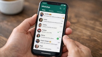 Primer plano de unas manos sosteniendo un smartphone negro con la aplicación de WhatsApp abierta, mostrando una lista de chats con nombres y mensajes en español.