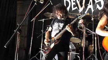 Un músico con cabello rizado y camiseta negra tocando una guitarra eléctrica en un escenario. Detrás, una batería, parte de otro músico y un letrero con la palabra 'DEVAS'