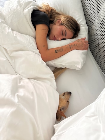 Una mujer con cabello claro duerme en una cama bajo sábanas blancas, con un brazo tatuado sobre una almohada, y un perro marrón claro asoma por debajo de la cobija