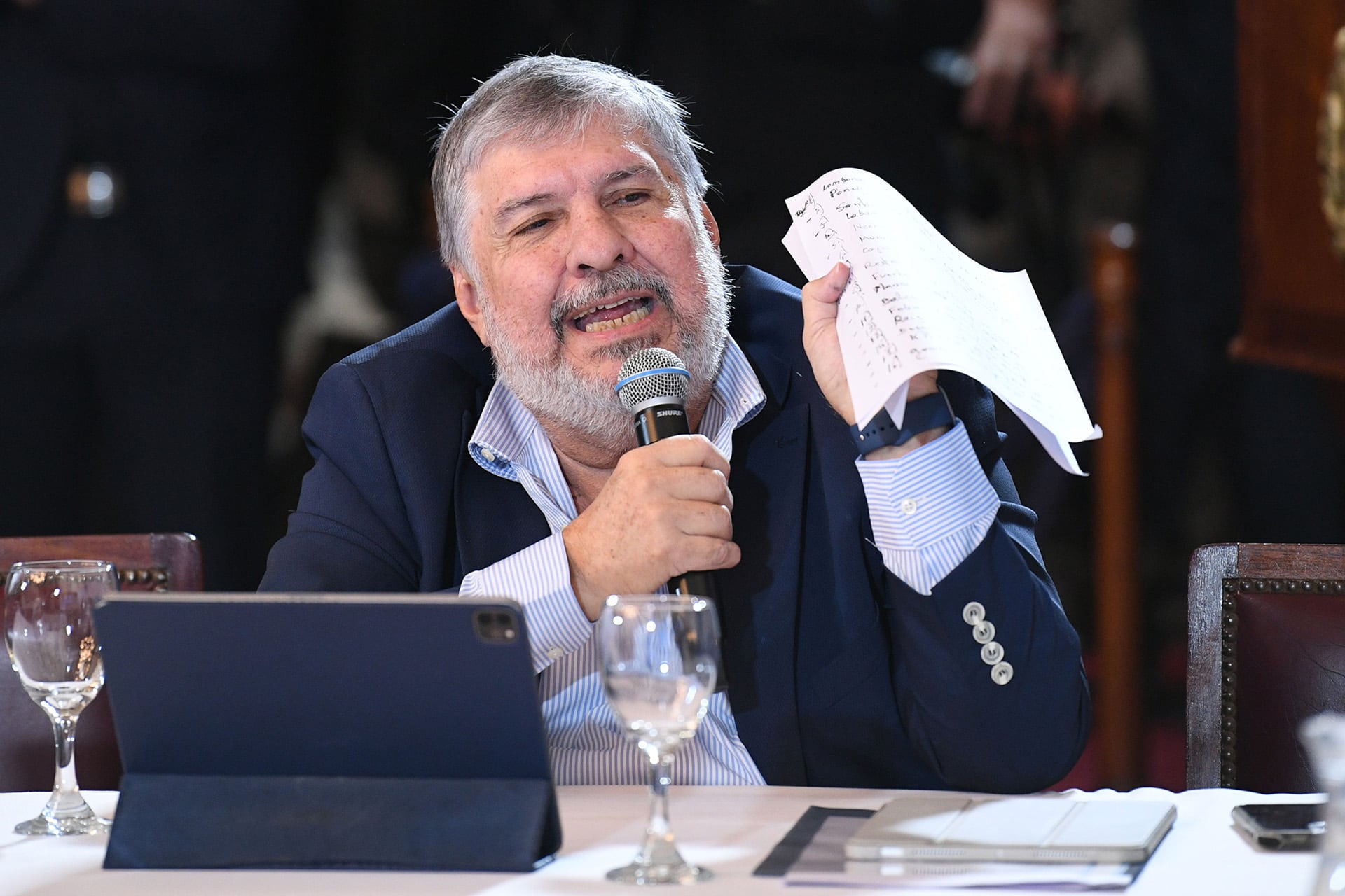 Un hombre con barba gris habla en un micrófono, sosteniendo una hoja de papel con notas, durante una presentación en un evento con una tableta y un vaso de agua en la mesa. (Mario Mosca/Comunicación Senado)