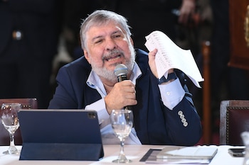 Hombre de barba gris y traje azul habla en micrófono, sosteniendo papeles. Hay una tableta y un vaso de agua sobre la mesa frente a él