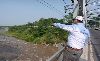 Hombre con camisa blanca de INSIVUMEH y gorra blanca apuntando hacia un río caudaloso y vegetación densa desde un puente con líneas eléctricas aéreas