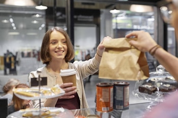 Una empleada sonriente con cabello corto entrega una bolsa de papel marrón y un vaso de café desechable a un cliente en un mostrador de tienda