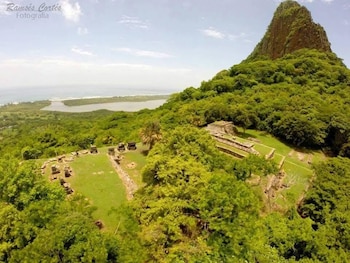Vista aérea de ruinas arqueológicas rodeadas de vegetación y una montaña rocosa; al fondo se observa una laguna y el océano bajo un cielo nublado