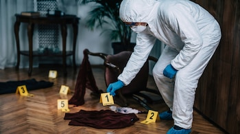Un experto forense con traje protector, mascarilla y guantes azules, coloca un marcador de evidencia numerado en un suelo de madera. Se observan más marcadores y objetos.