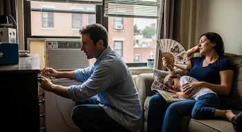 Un hombre de camisa azul se agacha para ajustar un aire acondicionado de ventana. Una mujer se abanica sentada en un sofá con un niño durmiendo en su regazo.
