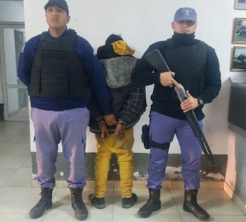 Dos policías uniformados, uno con escopeta, escoltan a un hombre con la cabeza cubierta y esposado, de pie en un interior