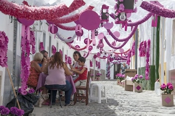 Varias personas sentadas a una mesa en una calle empedrada decorada con guirnaldas de papel y macetas de color rosa