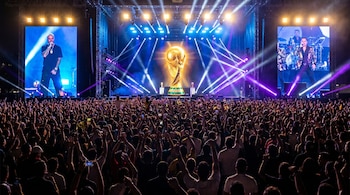 Gran multitud de fans con brazos en alto en evento nocturno. Escenario iluminado con trofeo Copa Mundial FIFA y artistas en pantallas.