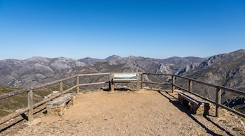 Mirador con suelo de tierra, valla de madera y dos bancos de piedra, con un cartel informativo y montañas de fondo bajo cielo azul.