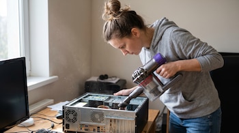 Una mujer con cabello recogido en un moño usa una pequeña aspiradora para limpiar el interior expuesto de una torre de computadora de escritorio.