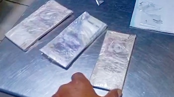 Tres fajos de billetes envueltos en plástico y varias bolsas pequeñas yacen sobre una mesa metálica junto a una mano