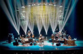 Cinco músicos en un escenario oscuro tocan instrumentos bajo intensos focos de luz blanca y azul. Una proyección de paisaje rocoso se ve al fondo
