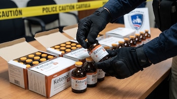 Manos enguantadas sostienen un frasco de Ketonal. Varias cajas abiertas y frascos de Ketonal se disponen sobre una mesa. Cinta amarilla de "POLICE CAUTION" en el fondo.
