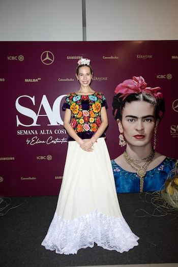 Una mujer con blusa bordada con flores y falda blanca posa frente a un panel morado con el retrato de Frida Kahlo y logotipos patrocinadores