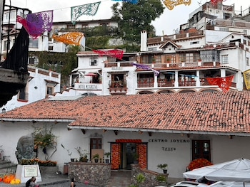 Vista panorámica de Taxco: edificios blancos con tejados rojos en una colina, decorados con papel picado, busto de bronce, personas y un coche