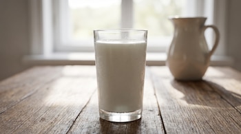 Un vaso de vidrio con leche fría y gotas de condensación sobre una mesa de madera, junto a una jarra de cerámica blanca y una ventana.