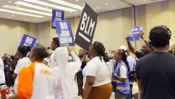 Varias personas con diversos tonos de piel sostienen carteles de protesta dentro de un gran salón. Los carteles visibles dicen "NO JIM CROW" y "BLM"