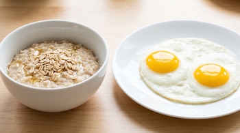 Un tazón blanco con avena y un plato blanco con dos huevos fritos reposan sobre una mesa de madera clara.