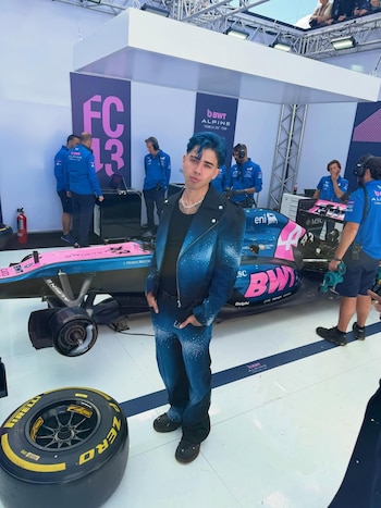 Luck Ra con cabello azul posa frente a un auto de Fórmula 1 Alpine rosa y azul, con un neumático Pirelli en primer plano y personal del equipo al fondo