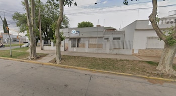 Edificio gris de una planta con el cartel "Dispensario 2", rodeado por una verja negra, árboles y una acera. Se ve una calle y casas al fondo