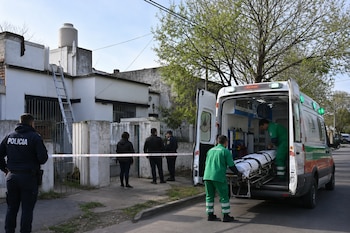 Una ambulancia abierta, personal de emergencia con una camilla, un policía y una casa con una escalera apoyada en el techo.