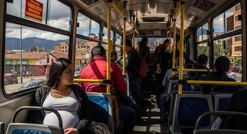 Vista interior de un autobús lleno de pasajeros en Bogotá. Una mujer embarazada sentada en primer plano mira hacia la ventana, revelando edificios y montañas.