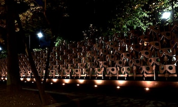 Vista nocturna de una pared decorativa de metal con formas geométricas en relieve, iluminada desde abajo por luces cálidas, con árboles y farolas al fondo