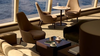 Salón de lujo con grandes ventanales con vistas al mar, sillones beige, mesas de centro con tazas de café y un periódico. Iluminación cálida y alfombra geométrica