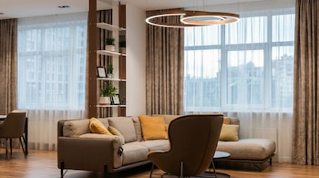 Sala moderna con sofá esquinero beige, sillas marrones, estantería, lámpara circular y ventanas grandes.