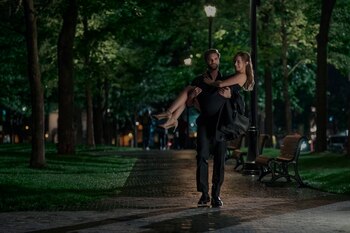 Hombre barbudo con ropa oscura carga a mujer rubia de vestido negro y tacones en un camino adoquinado por la noche, rodeados de árboles y farolas