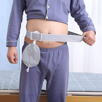 Torso e pernas de homem de pijama azul com cós elástico e bolsa coletora cinza. Sua mão ajusta o cinto. Cama nos fundos