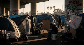 Vista de un campamento improvisado bajo un paso elevado de una autopista en Los Ángeles, con varias tiendas de campaña, personas y pertenencias al atardecer.