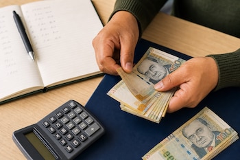 Manos de una persona contando billetes de cien soles peruanos junto a una calculadora, un cuaderno abierto y un bolígrafo en una mesa.