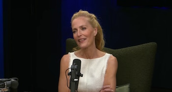 Una mujer rubia de mediana edad sonríe, vestida con un top blanco sin mangas, sentada. Un micrófono negro se ubica frente a ella. El fondo es oscuro