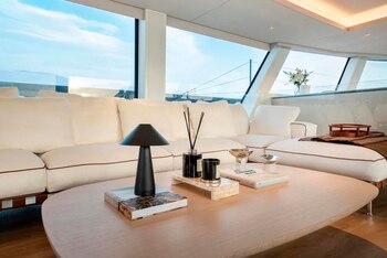 Salón con un sofá blanco y una mesa de madera con lámpara, difusor y libros frente a amplios ventanales que dan al exterior