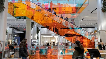 Vista interior de un centro comercial moderno con una escalera mecánica de color naranja brillante. Se ven personas subiendo y bajando, y tiendas de lujo