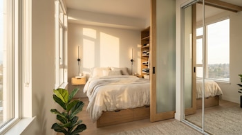Dormitorio moderno con cama beige, mesita de noche flotante, estantería de madera y grandes ventanales. Una planta verde en primer plano y puertas corredizas.