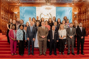 Un grupo diverso de hombres y mujeres posa en una escalera alfombrada de rojo, con la bandera de Guatemala de fondo y paneles de madera en las paredes